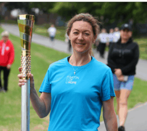 A person wearing a blue 'BATON OF HOPE' t-shirt holds a baton with a golden top while standing on a pathway outdoors. Several people are walking in the background.