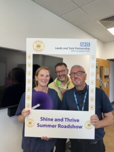 A photo on the right side shows three people holding a large frame that reads: "Shine and Thrive Summer Roadshow" The frame includes logos for Leeds and York Partnership NHS Foundation Trust.