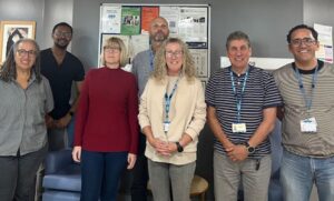 Seven people standing in front of a bulletin board with posters and notices. They are casually dressed, some wearing ID badge lanyards. Chairs and a framed picture are visible in the background