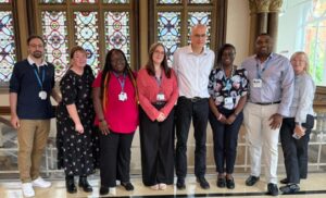 A group of eight individuals standing in a line indoors, dressed in business casual clothing with visible ID badges. Behind them are ornate stained glass windows and a marble floor.
