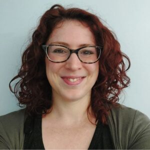 A person with dark curly hair smiles at the camera, she is wearing glasses. 