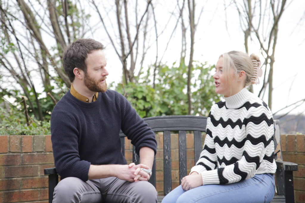 Two people sit on a bench facing each other in conversation.