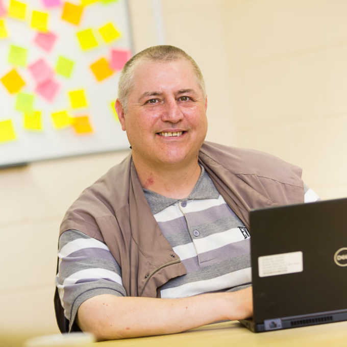 Paul Fraser is sat at a desk in front of an open laptop. He is smiling towards the camera. In the background there is a whiteboard with lots of brightly coloured sticky notes.
