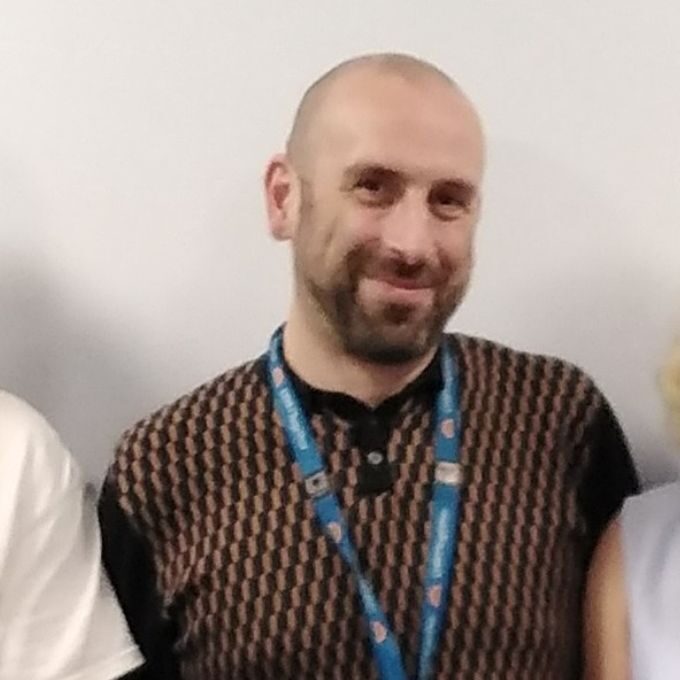 A medium close up of Matt. Two people are stood either side of him but they are cut out of the image. Matt is wearing a blue lanyard and patterned top