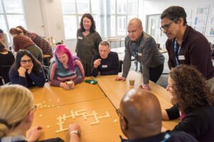 A diverse group of people gathered around a table, intently playing a game of Scrabble with tiles and a game board