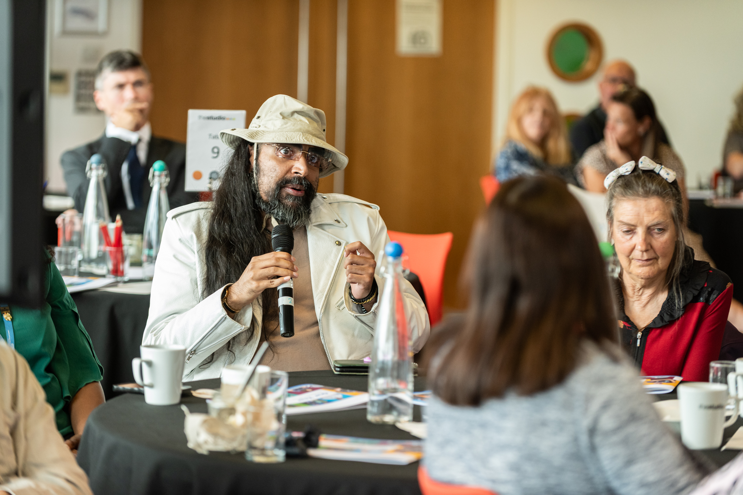 A gentleman with a beige hat, jacket and beard speaks into a microphone seated at a table