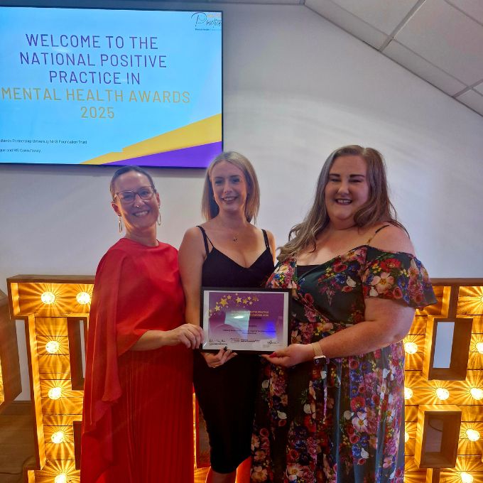 Three individuals standing indoors holding an award plaque at the National Positive Practice in Mental Health Awards 2025. A welcome sign and decorative lighting are visible in the background.