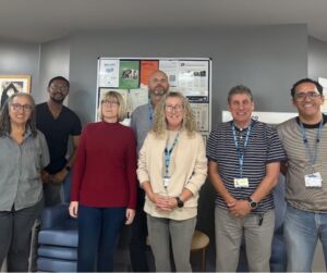 A group of six people standing and smiling in a hallway.