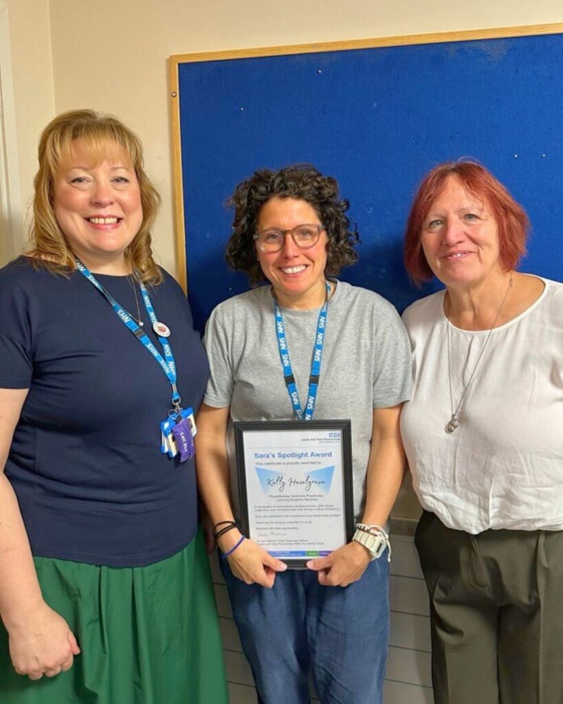 Photograph of three people standing smiling at the camera. The lady in the middle is holding a certificate.