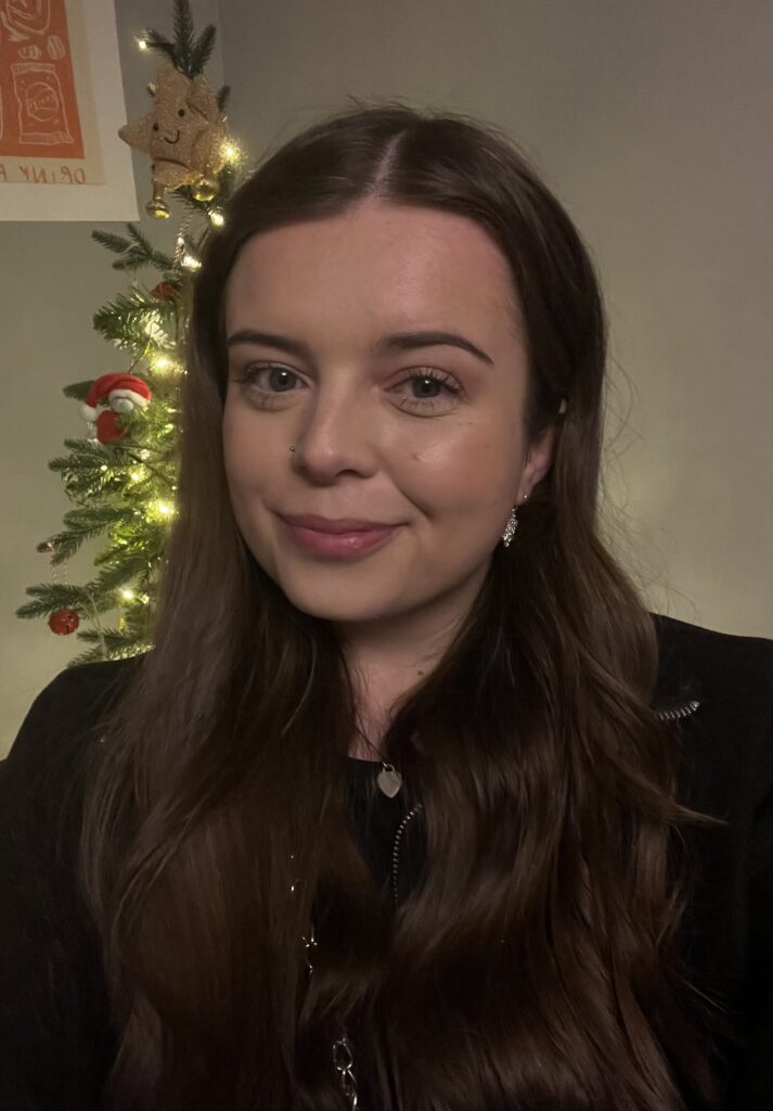 A photograph of Charlotte Kelly, she has long dark brown hair, is wearing a black shirt and is smiling at the camera.