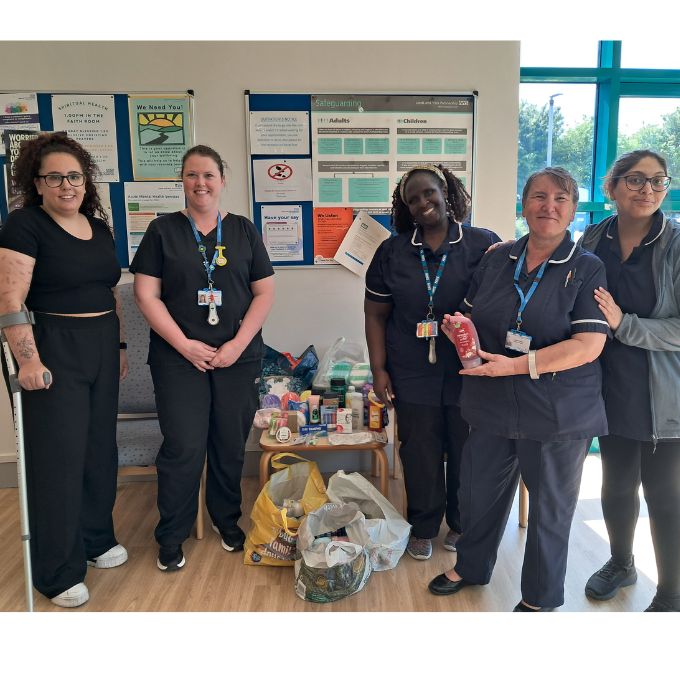 Healthcare staff members and Chloe are standing indoors next to a small table filled with donated items. The table holds various goods including canned food, toiletries, and household supplies, with additional bags of donations placed on the floor in front. Behind them is a noticeboard displaying multiple posters and information sheets. A large window on the right side lets in natural light.