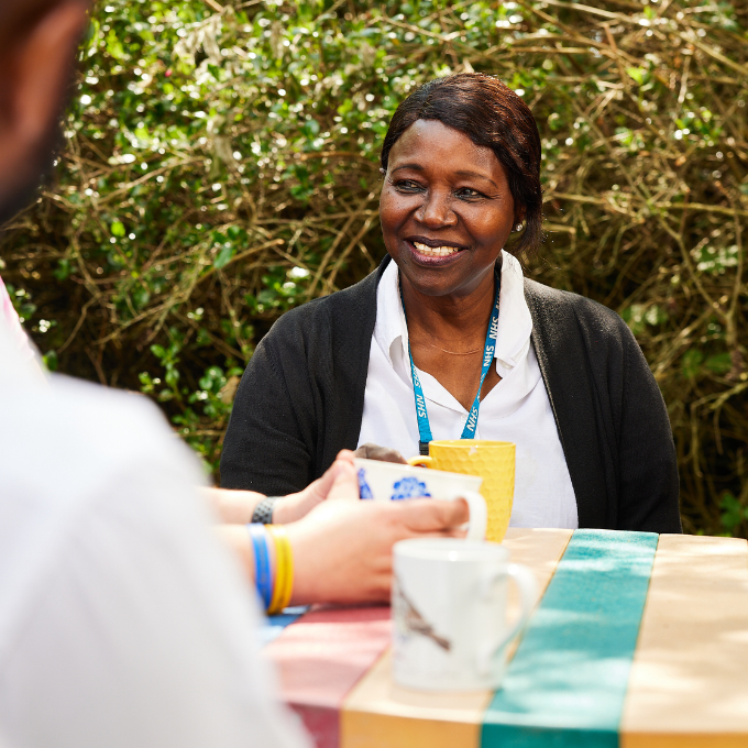A member of staff with an NHS lanyard, white shirt, and black cardigan, sitting at a colourful table having a cup of tea with colleagues