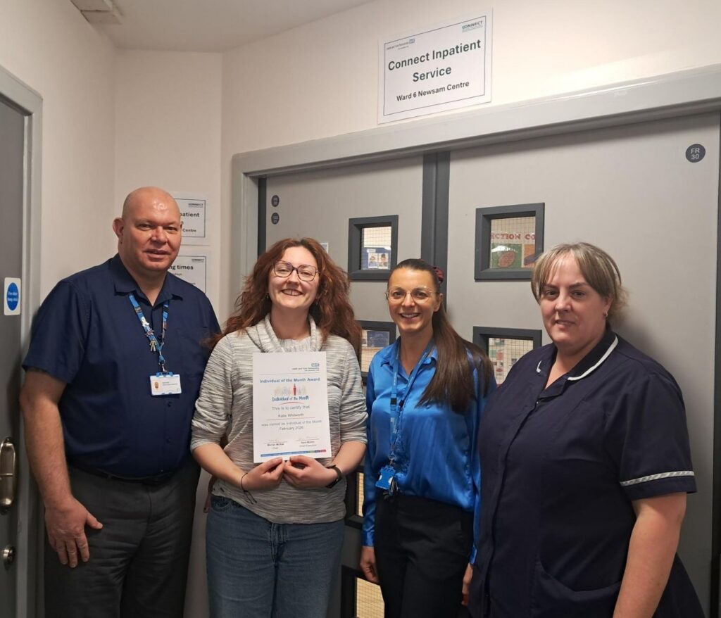 A group of four people standing in front of grey double doors. they are all looking at the camera and smiling. One person is holding a certificate.