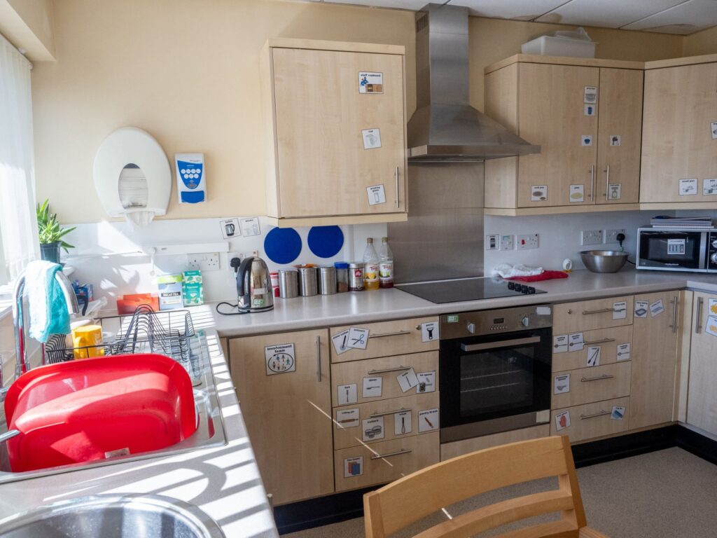 A photograph of an accessible kitchen. There are easy read icons on the cupboards and clear notices around. 