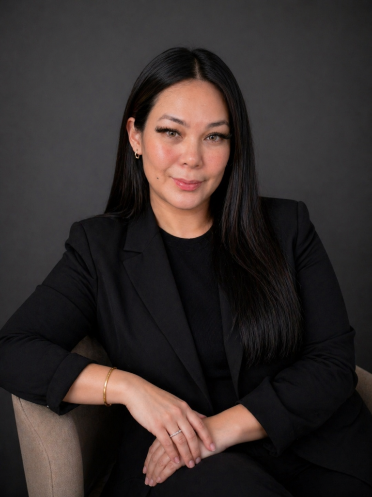 A woman in a black blazer smiles gently at the camera, seated against a dark grey background.