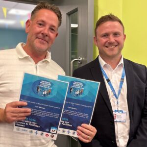 One man proudly holds 2 certificates while another stands beside him in a room setting. Both are smiling