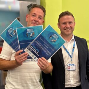 One man proudly holds 3 certificates while another stands beside him in a room setting. Both are smiling