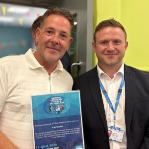 One man proudly holds a certificate while another stands beside him in a room setting. Both are smiling