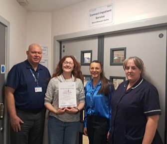 Four people pose for a photo, some in nurse uniforms. One person holds a certificate.