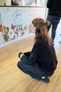 A woman crouching to look at a wall-mounted display board covered in notes and botanical collages.