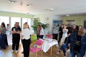 A diverse group of people socialising in a bright, informal communal room with colourful chairs and a notice board.