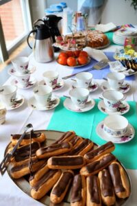 A table set for tea with a platter of chocolate eclairs, vintage floral teacups, coffee carafes, and assorted cakes.