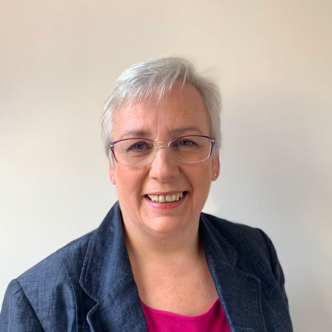A lady with glasses, blue jacket and pink top smiling at the camera.