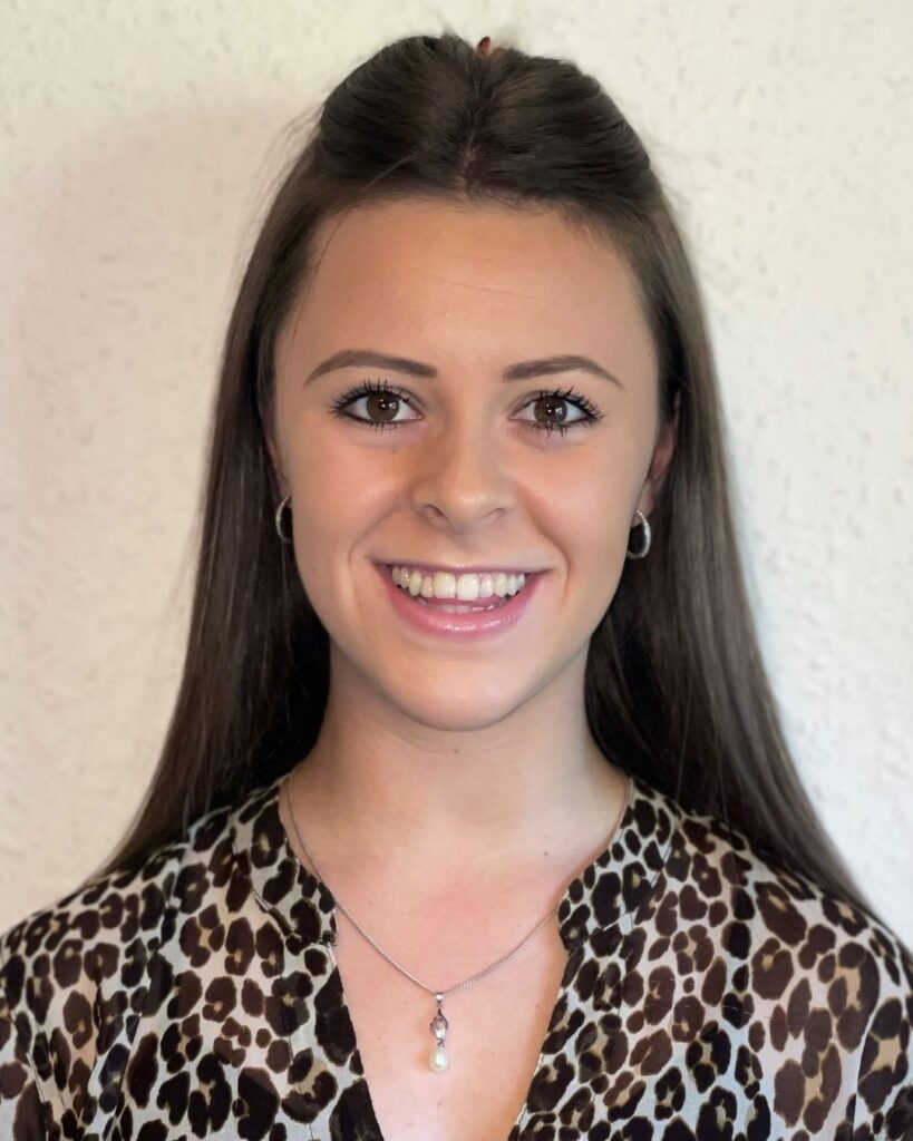 A smiling young woman with long brown hair, wearing a leopard print top and a pearl pendant necklace.