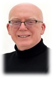 Head-and-shoulders studio portrait of a person wearing black rectangular glasses and a black ribbed turtleneck sweater. The person has a shaved or bald head and is facing the camera directly. The image is set against a plain light grey background.