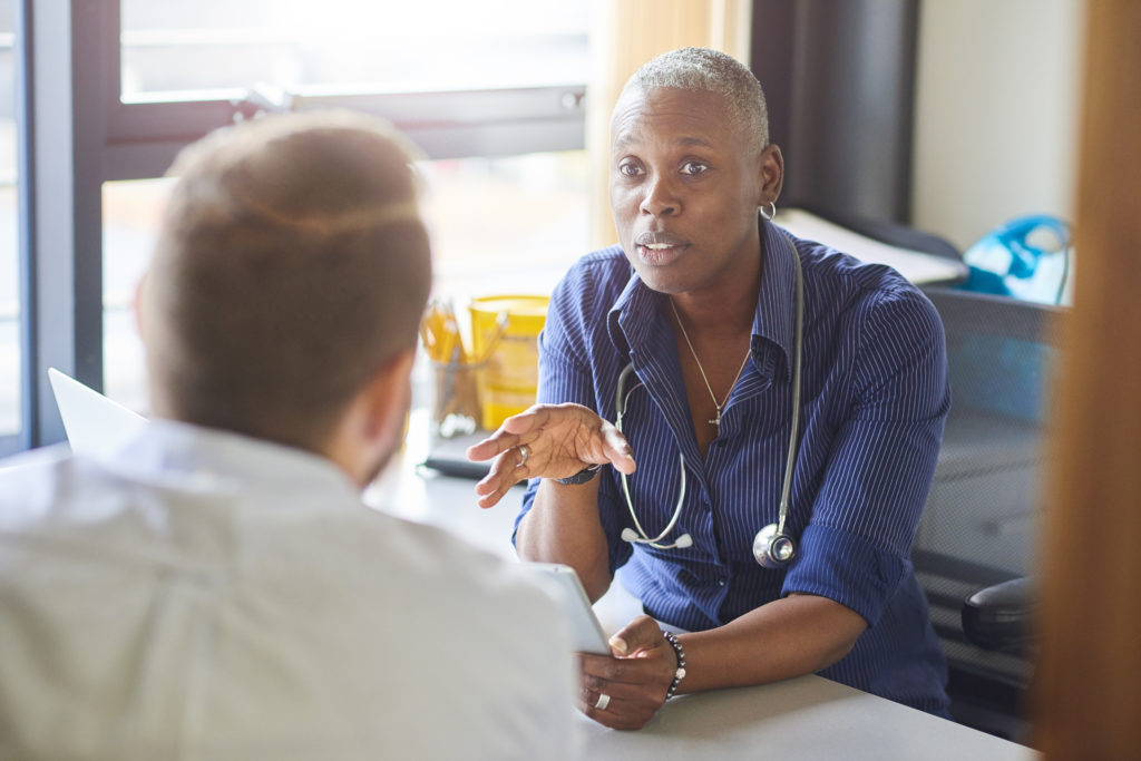 A doctor sits at her desk and chats to a male patient.