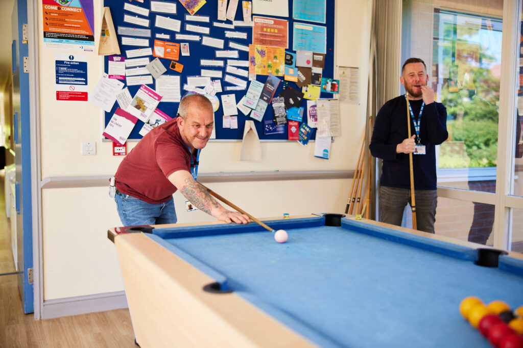 Two people are playing pool. One person is bent over the table holding a pool cue which is poised against the white ball ready to shoot. 