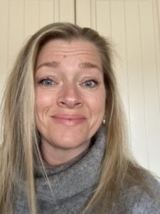 Tania is wearing a grey turtle neck jumper and smiling. Tania has long blonde hair.