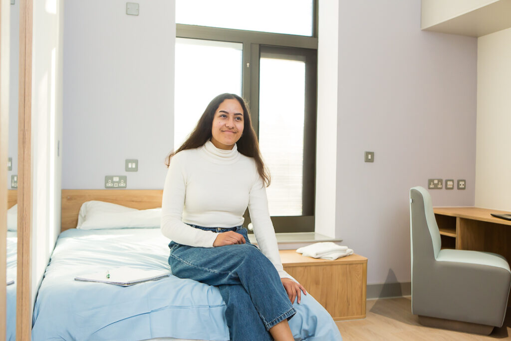 A young person is sat on the end of a single bed with blue duvet. The room has a large window and a desk.