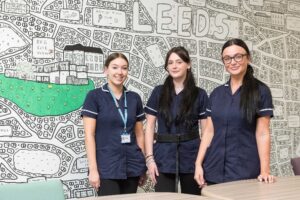 Three staff members in navy uniforms standing together in front of a large illustrated wall map featuring the word ‘Leeds’ and hand‑drawn buildings and streets.