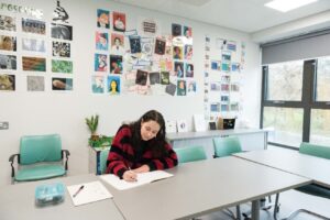 A person sitting at a table in a bright classroom, writing in a notebook. The walls are decorated with colourful artwork and photo displays, and large windows let in natural light.