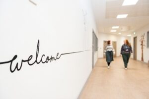 A bright corridor with the word ‘welcome’ written in script on the wall, and two people walking together further down the hallway.