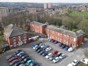 Aerial view of the St Mary’s Hospital site, showing several connected red‑brick buildings arranged around a large central car park filled with vehicles. Surrounding the site are tall residential blocks, houses, trees and green spaces stretching into the distance.