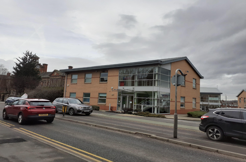 Two-storey modern brick building with a glass-fronted entrance and upper-floor windows, viewed from across a road with several parked and passing cars under an overcast sky.