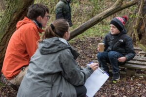 Two adults are kneeling in front of a child who is sat on a log in a wood.