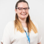 A medium close up of a young women smiling towards the camera. She is wearing glasses, a white top and blue lanyard.