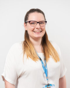 A medium close up of a young women smiling towards the camera. She is wearing glasses, a white top and blue lanyard.