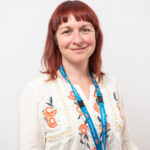 A person with shoulder‑length red hair is wearing a light cream blouse decorated with orange and dark blue floral embroidery. The person has a blue LYPFT lanyard around their neck and is standing against a plain white background.