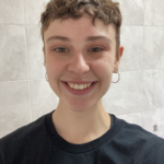 A person with short, wavy hair is shown in a close-up photo, facing the camera. The person is wearing small hoop earrings and a dark T‑shirt. The background consists of light-coloured tiled walls