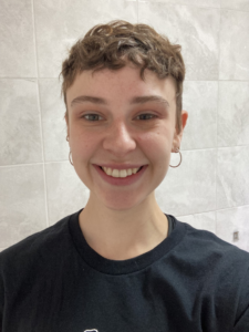 A person with short, wavy hair is shown in a close-up photo, facing the camera. The person is wearing small hoop earrings and a dark T‑shirt. The background consists of light-coloured tiled walls