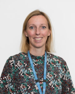 A person with light‑coloured, shoulder‑length hair is wearing a high‑neck, long‑sleeved top with a dense floral pattern in green, white, red and black. A blue LYPFT lanyard hangs around the person’s neck. The background is plain and white.