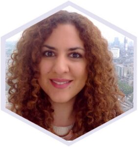 A smiling person with long, curly light brown hair is looking directly at the camera. They are wearing a light-coloured knit top and a statement necklace. A city skyline is visible through a window in the background on a cloudy day.