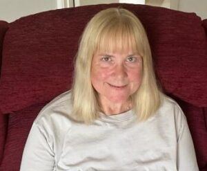 A close-up indoor photo of a person sitting on a red upholstered chair. The person has straight, light-coloured shoulder-length hair with a fringe and is wearing a plain light grey top. The background is neutral and softly lit, with the focus on the person facing the camera.