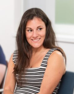 Person with long dark hair wearing a black-and-white striped sleeveless top, seated during a meeting or discussion.