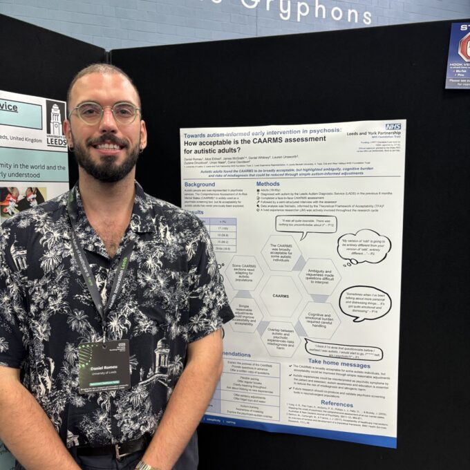 A person wearing glasses, a dark patterned shirt and a conference lanyard stands beside a research poster displayed at an academic event. The poster, affiliated with Leeds and York Partnership NHS Foundation Trust and the University of Leeds, explores the acceptability of the CAARMS assessment for autistic adults. The display is mounted on black exhibition boards, with the words “Home of the Gryphons” visible on the wall above.