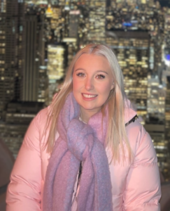 Portrait of a person wearing a light pink padded coat and a large lilac scarf, seated outdoors at night. The background shows a blurred city skyline with illuminated buildings and windows, creating a bokeh effect. The person is centred in the image and facing the camera.